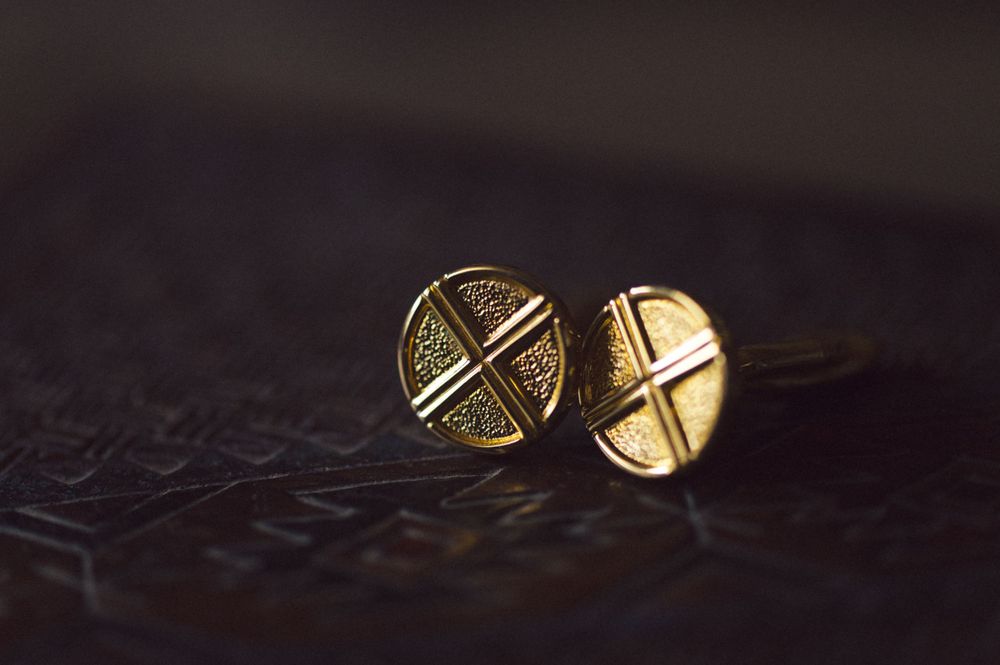 Two round gold cufflinks with crossed patterns photographed on a dark textured surface.
