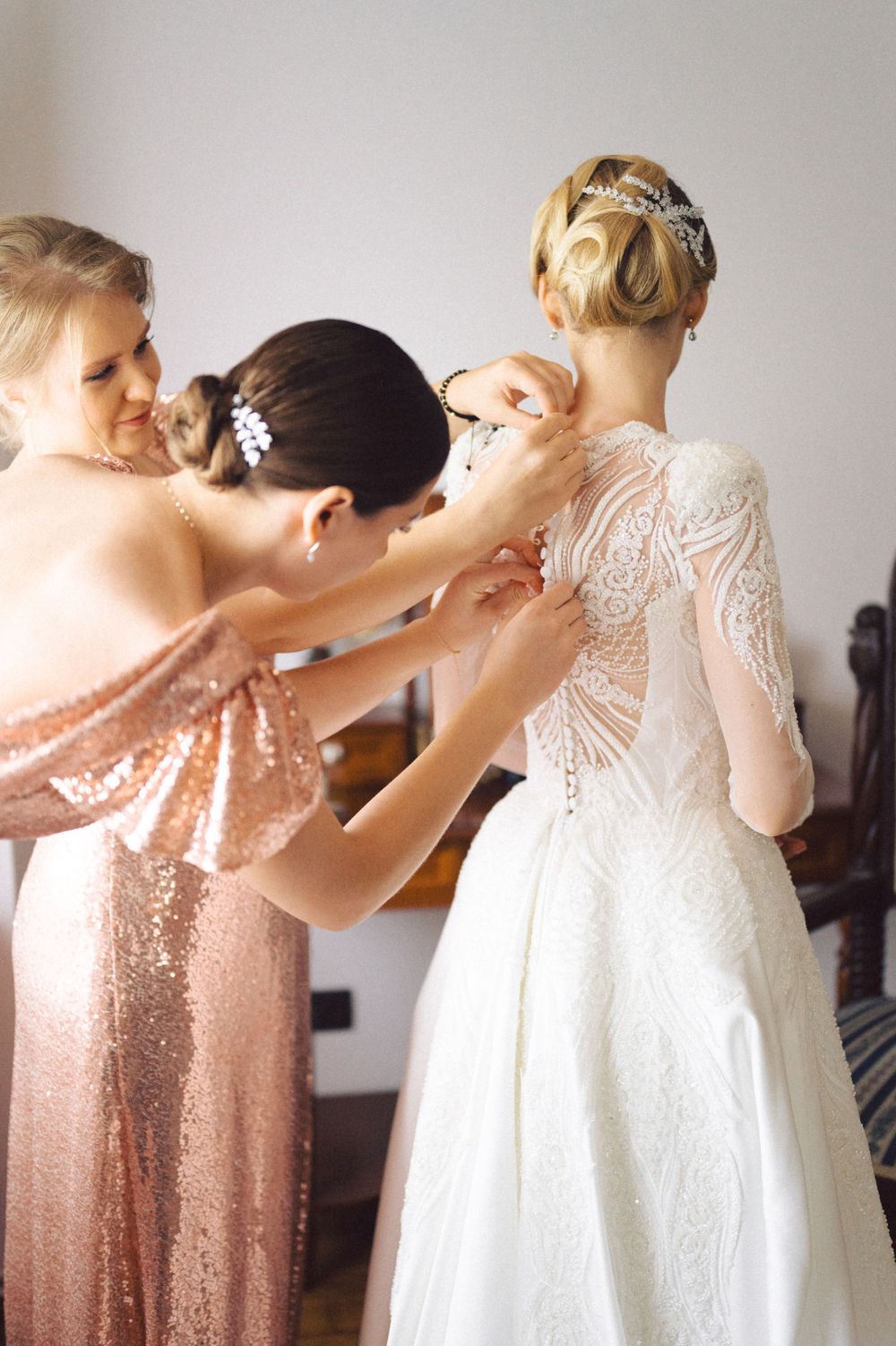 Bridesmaids in rose gold sequin dresses help adjust a wedding gown featuring delicate lace details.