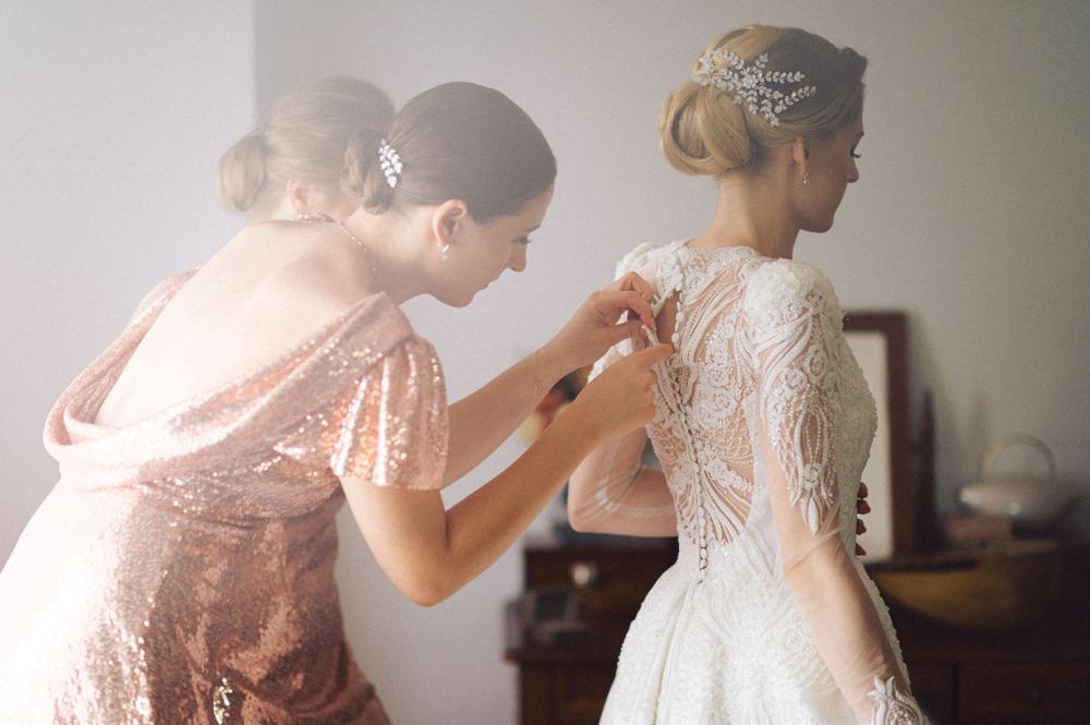 A bridesmaid in a sparkly rose gold dress helps secure the back of an elegant white lace wedding gown.