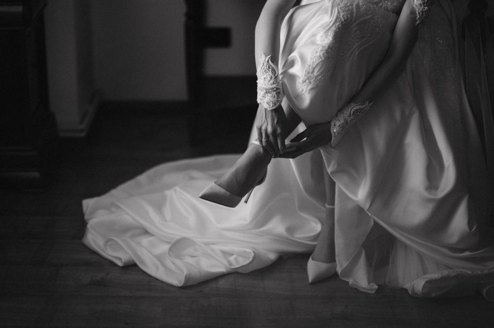 Black and white image of wedding dress and shoes being adjusted on wooden floor.