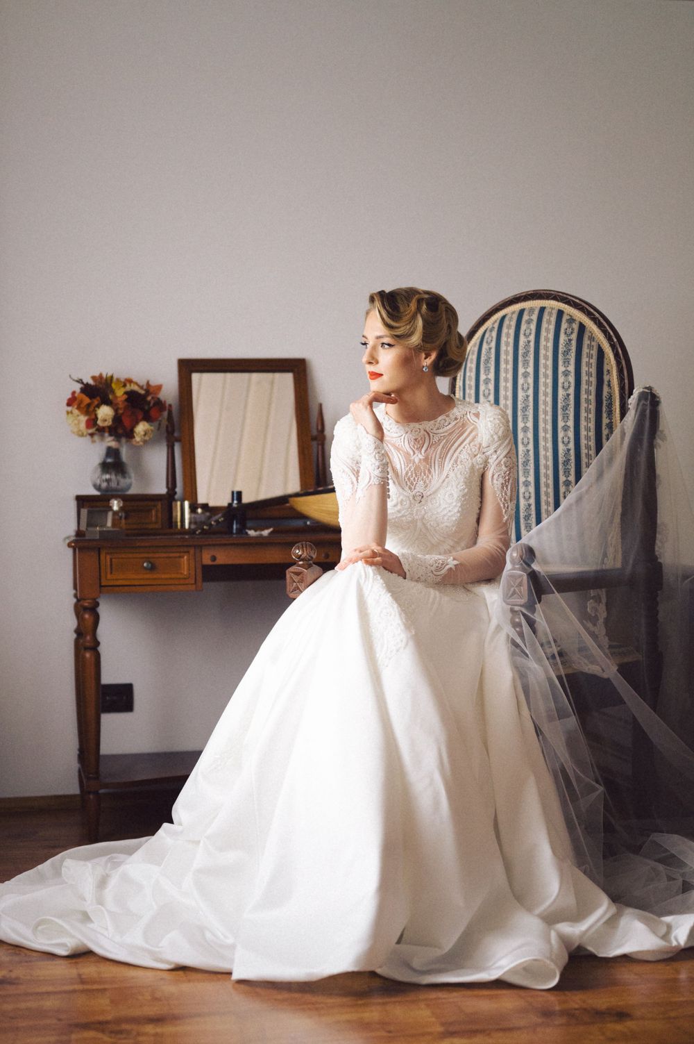 Elegant bride in vintage-inspired lace wedding gown sits at antique vanity with floral arrangement and mirror.
