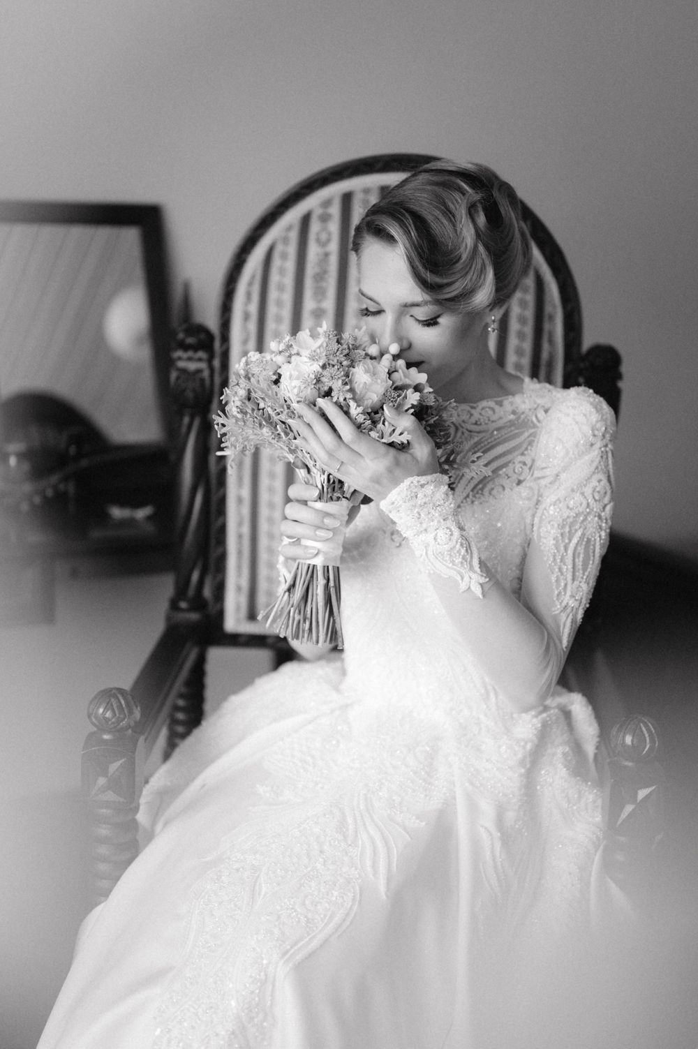 Black and white portrait of bride in lace gown holding bouquet while seated in vintage chair.