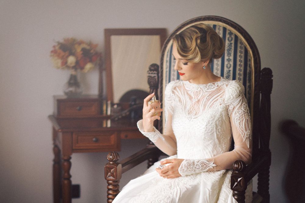 Bride in long-sleeved lace wedding dress sits in vintage wooden chair near antique vanity.