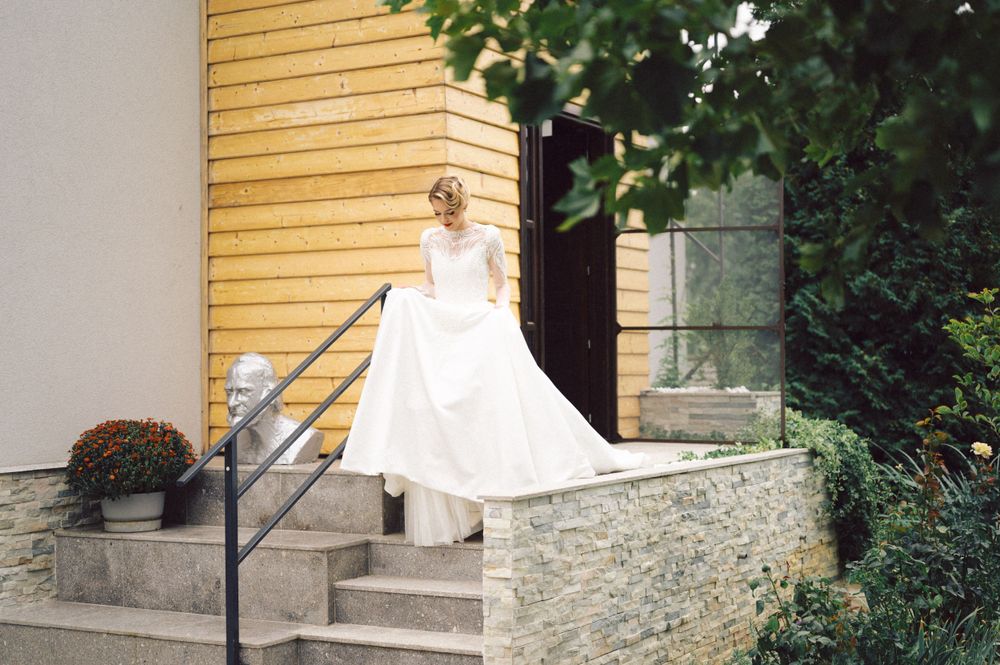 Bride in flowing white gown stands on stone steps outside yellow building with autumn decorations.