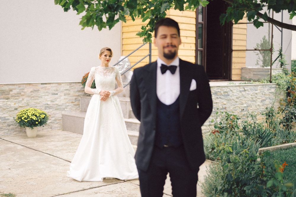 First look wedding photo with groom in tuxedo standing in foreground and bride on steps in background.