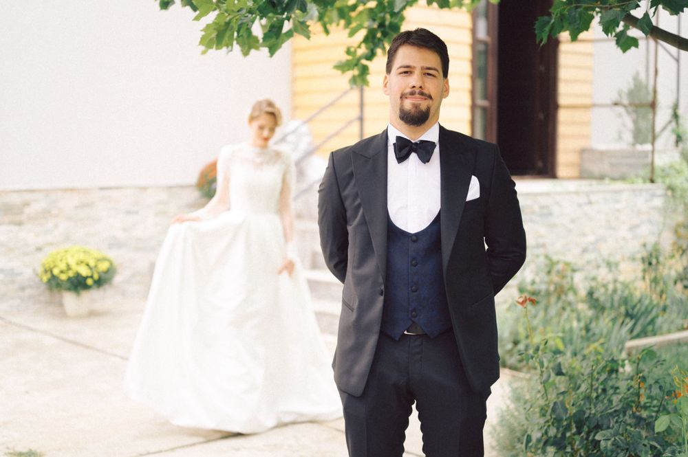 Romantic first look moment with groom smiling in black tuxedo while bride waits in background.