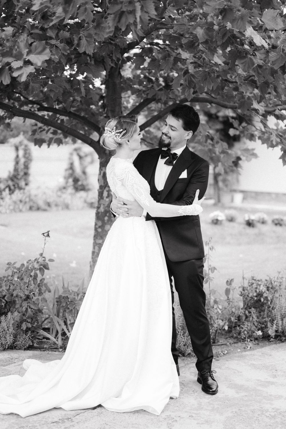 Elegant black and white wedding photo of a couple embracing under a tree in a garden setting.