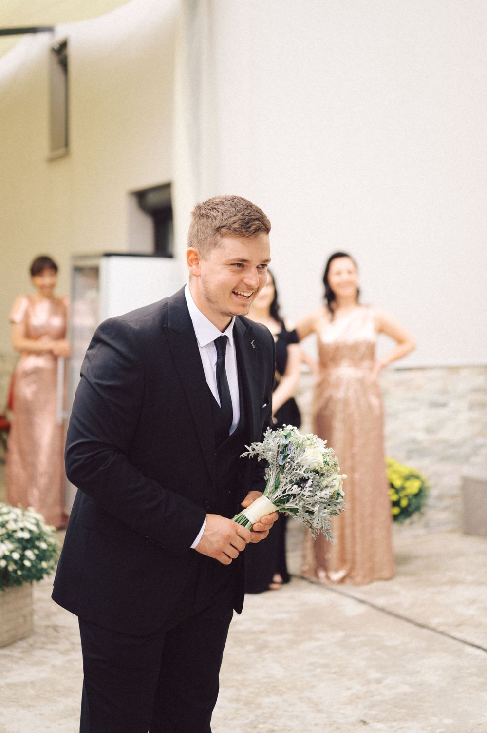 Smiling person in black suit holding small floral boutonniere outdoors.