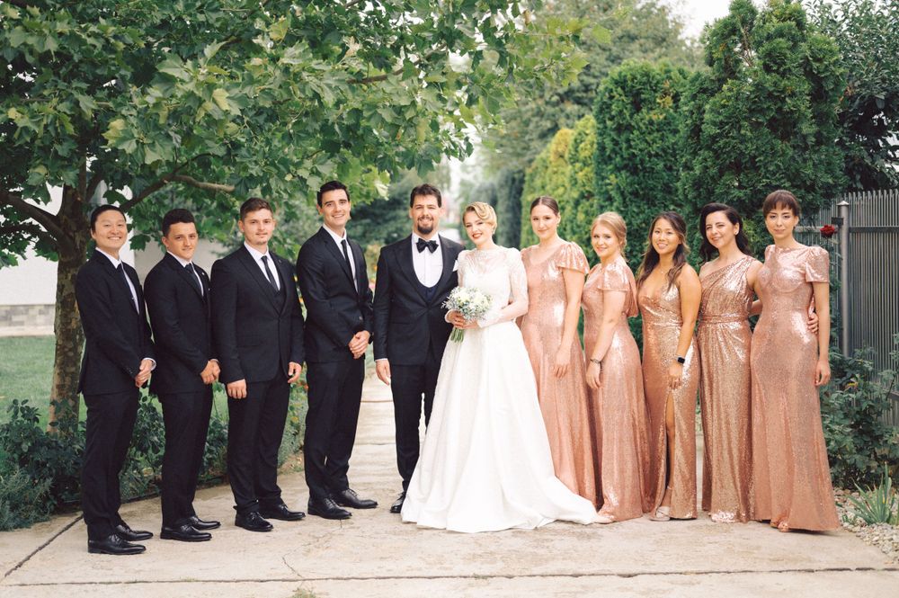 Wedding party in black suits and rose gold dresses posed together outdoors.
