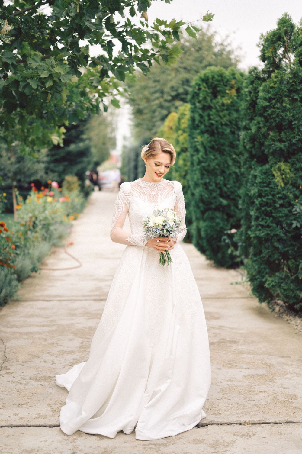 Elegant bride in white lace gown holding bouquet on garden pathway.