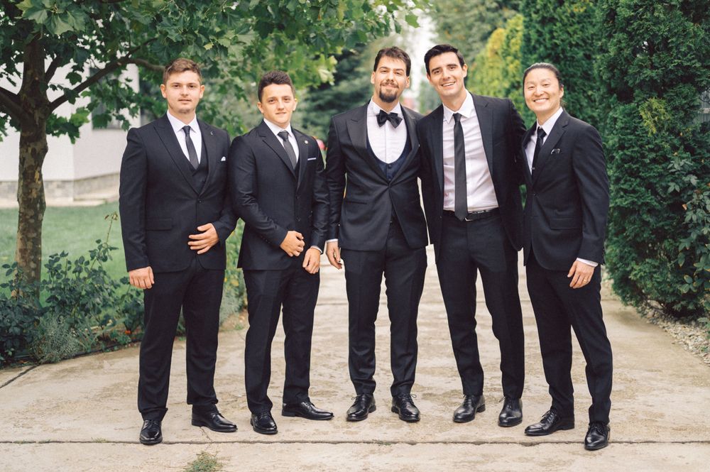 Five groomsmen in black suits posing together on garden path.