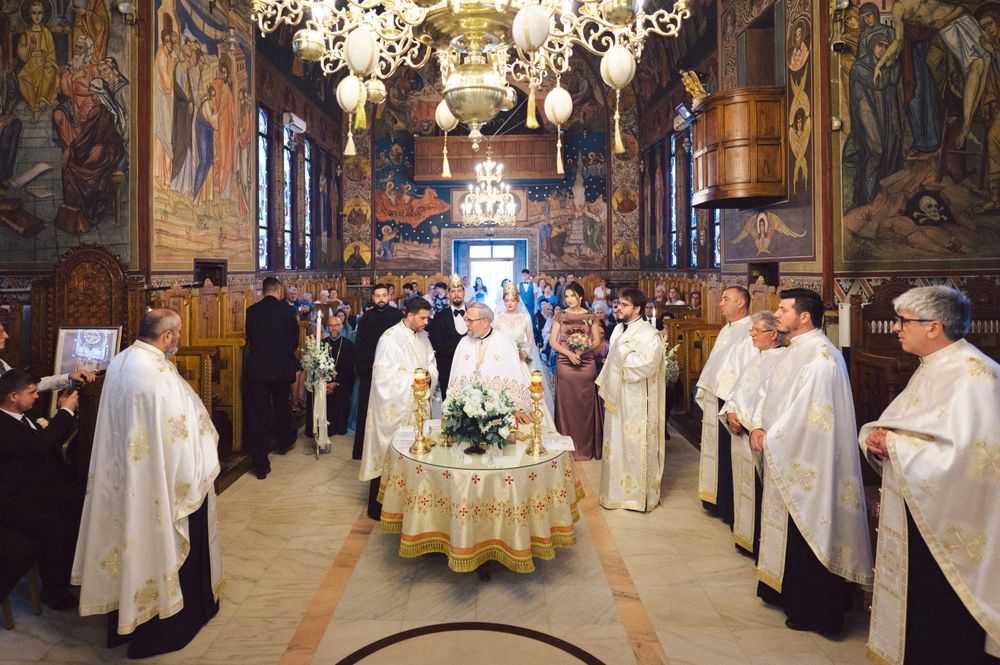 Orthodox Christian wedding ceremony in an ornate church with clergy in white robes and a grand chandelier overhead.