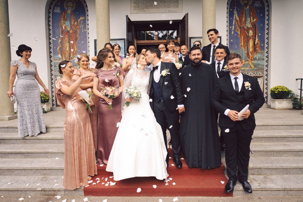 Wedding party poses on church steps with bridesmaids in rose gold dresses and clergy in black robes.