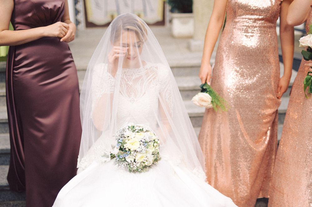 Bridesmaids in burgundy and rose gold dresses surrounding bride in white wedding gown with bouquet.