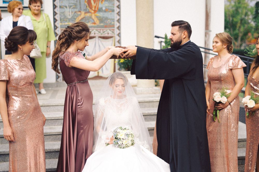 Wedding party gathered outside church with bridesmaids in metallic dresses and clergy in traditional robes.
