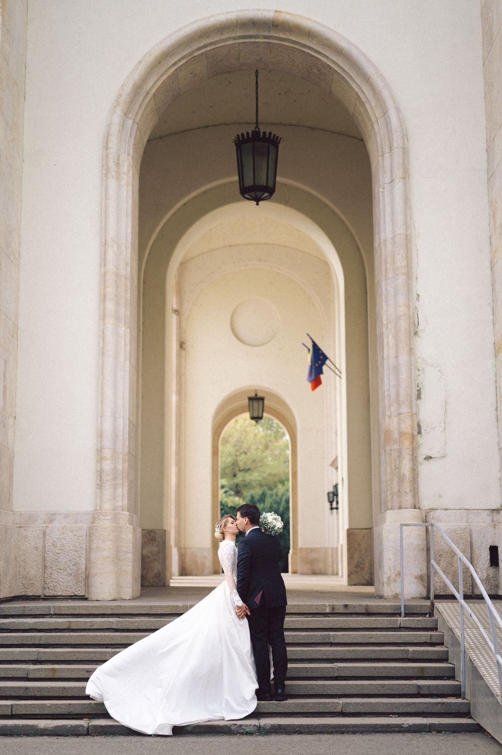 Newlyweds embrace under grand archway with white columns and French flag in elegant formal wear.