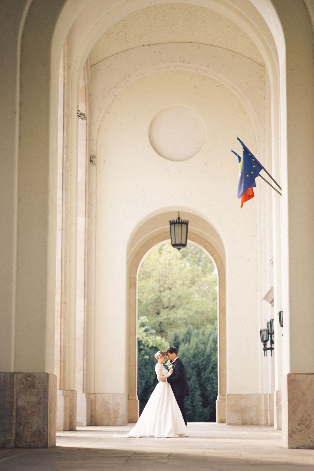 Wedding couple shares intimate moment beneath arched colonnade with French flag overhead.