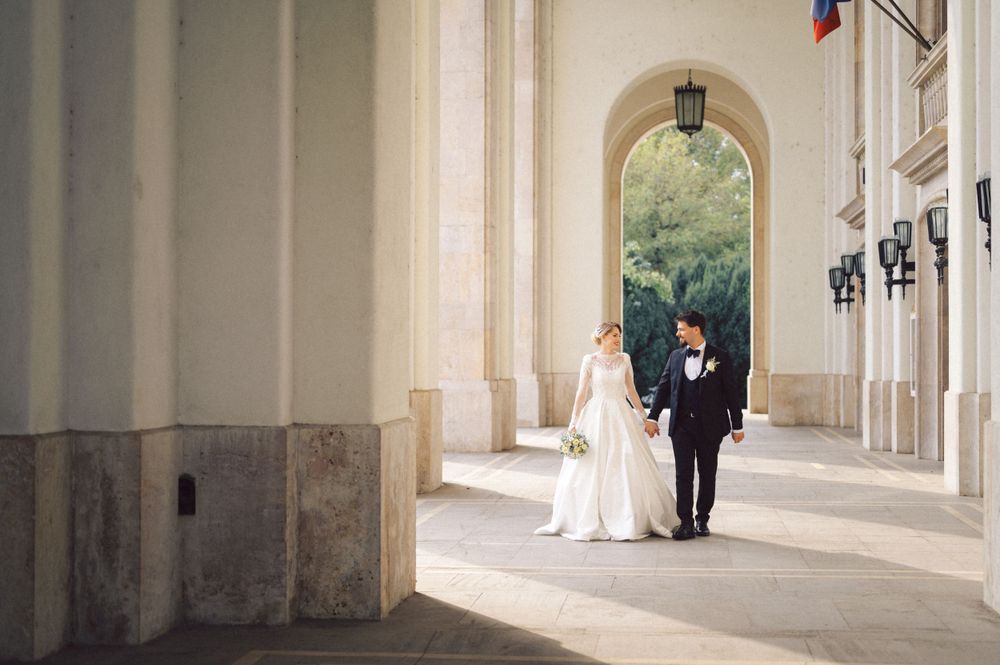 Bride in white gown and groom walk together under classical archway with hanging lanterns.