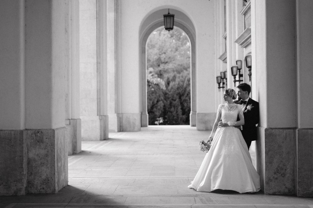 Black and white photo of wedding couple posing beneath ornate architectural arches.