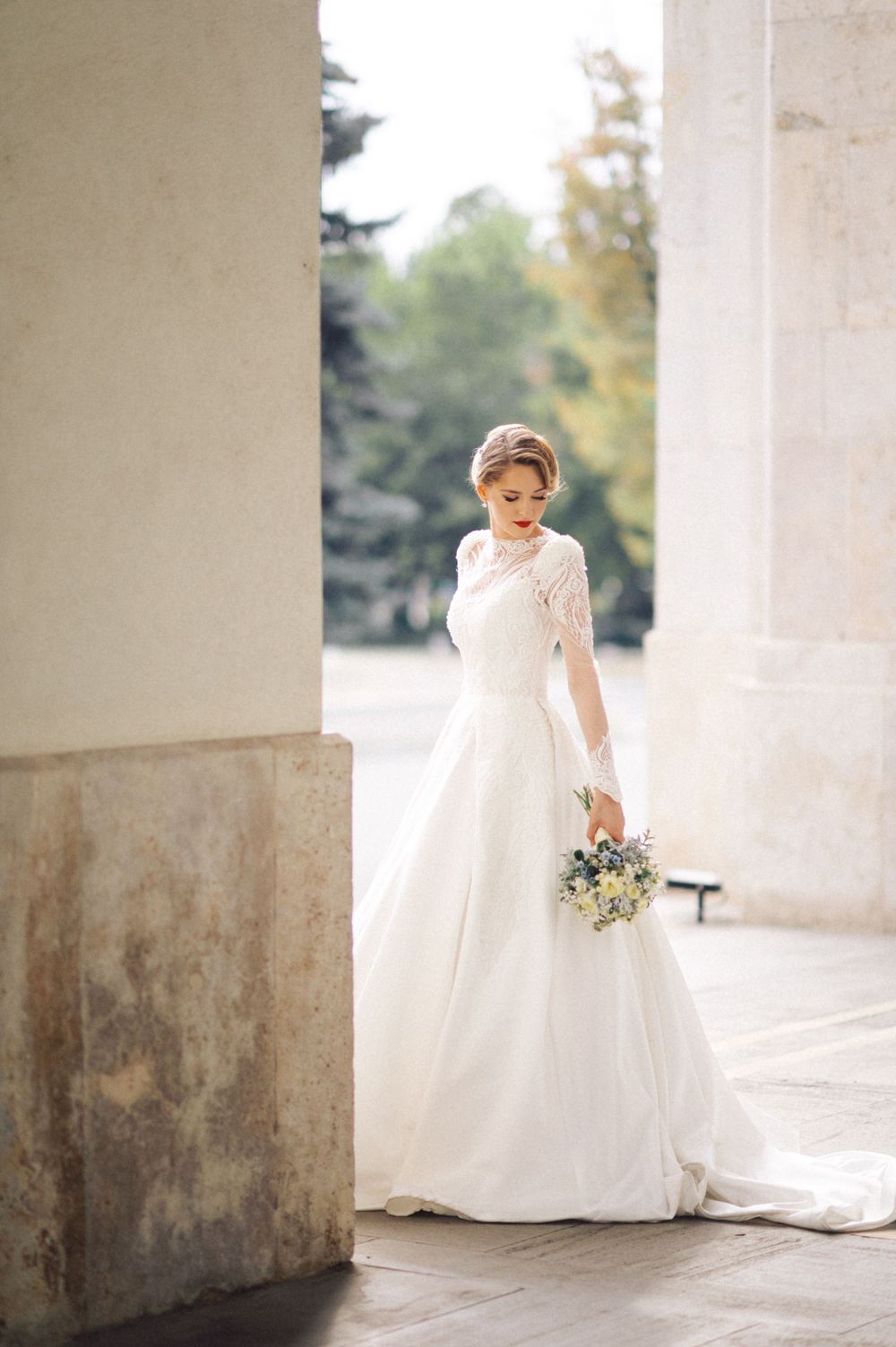 Elegant bride in lace-detailed wedding gown holds bouquet while posing by classical column.