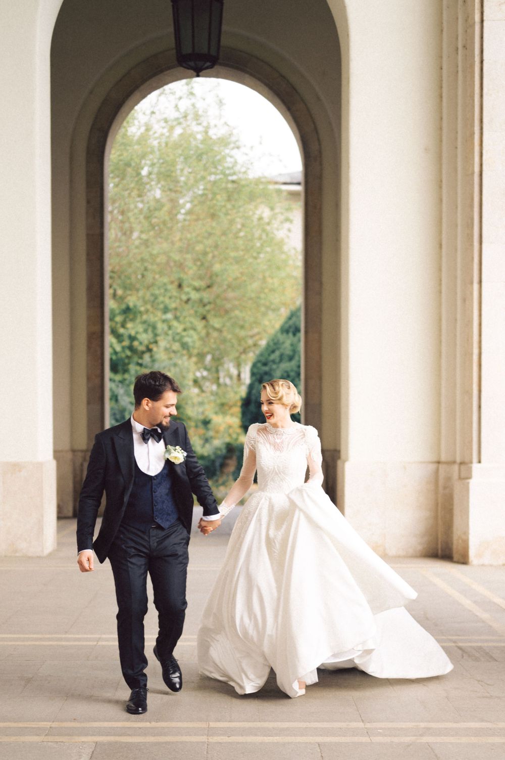 Wedding couple walks hand in hand through grand archway in formal attire.