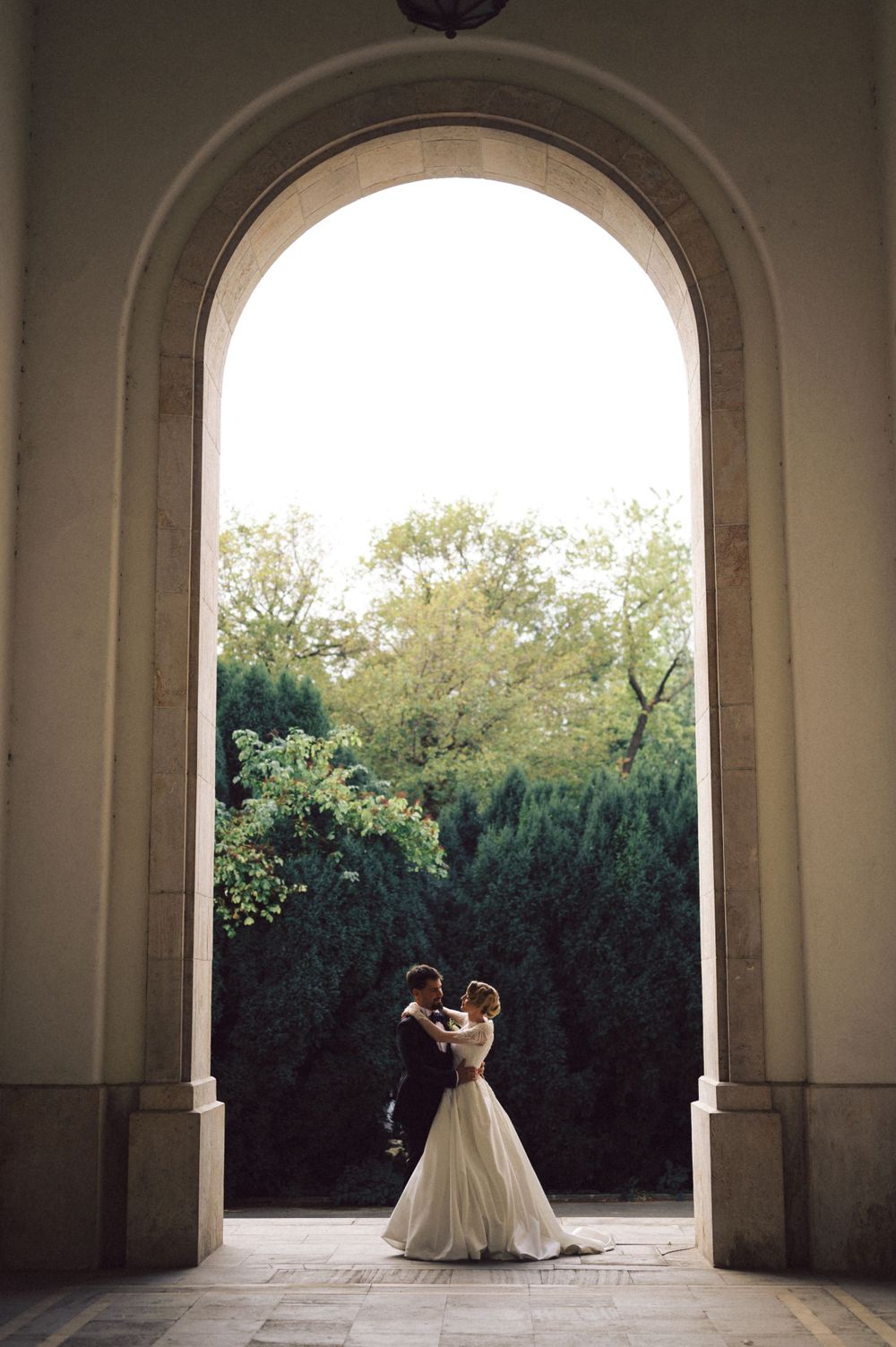 Silhouetted wedding couple embrace under stone archway framed by garden greenery.