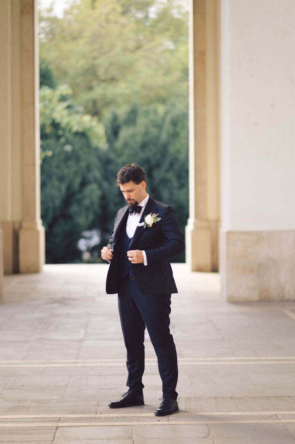 Groom in classic black tuxedo checking time while standing in elegant columned walkway.