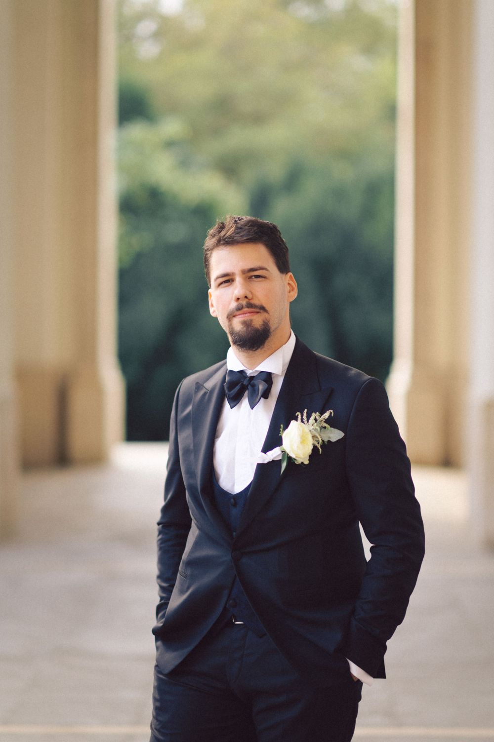 Classic portrait of groom in black tuxedo and bowtie standing in columned walkway.