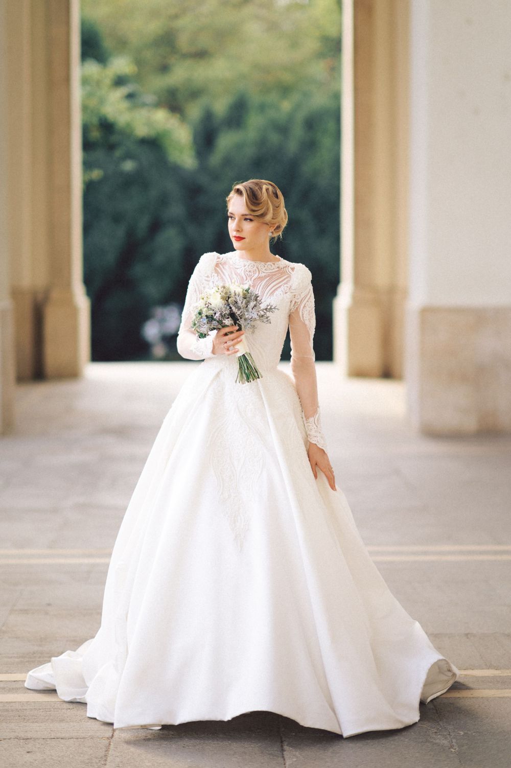 Elegant bride in long-sleeved white ballgown holding lavender bouquet in columned courtyard.