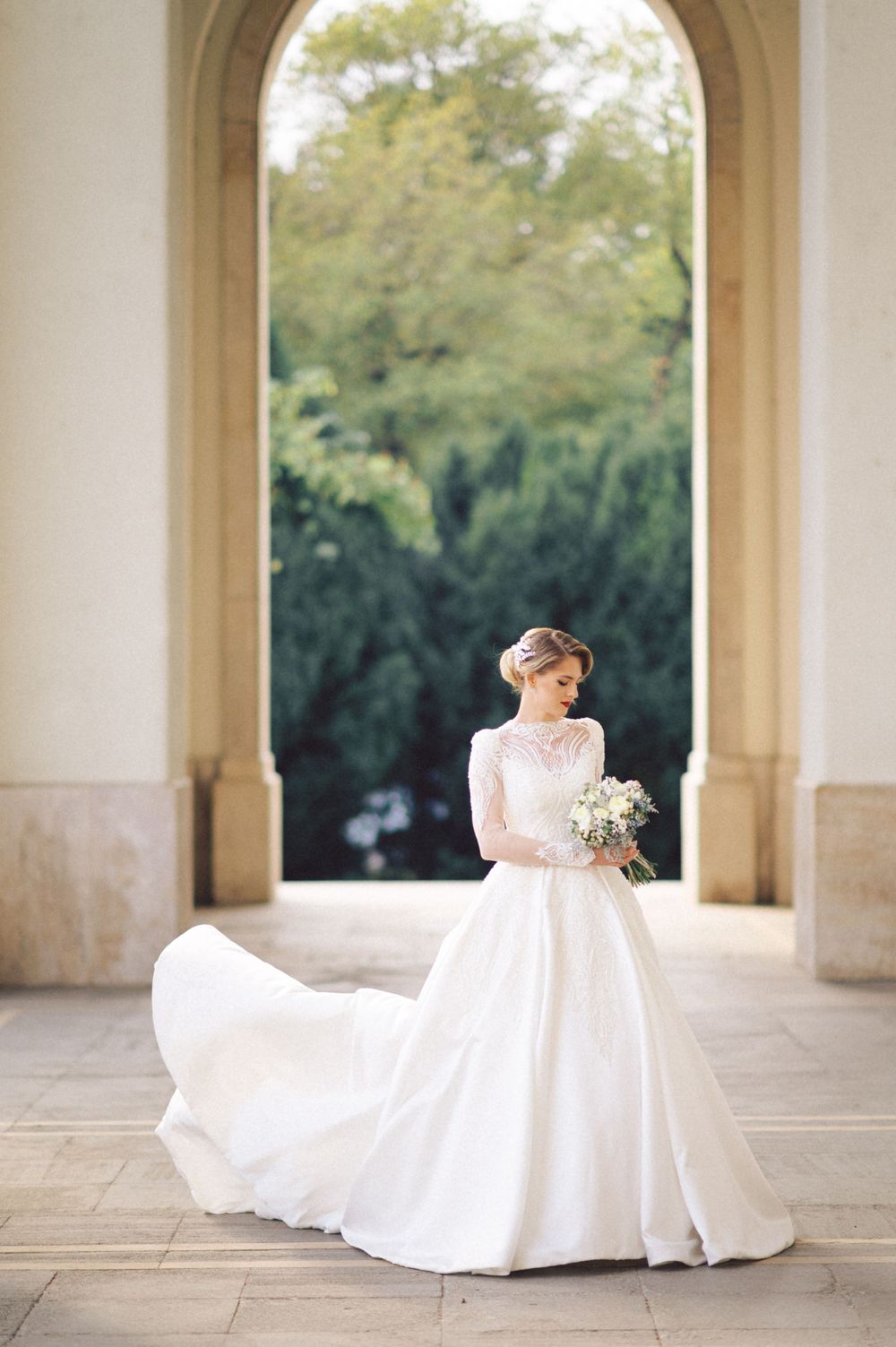 An elegant bride in a flowing white lace gown with long sleeves poses in a grand archway entrance.