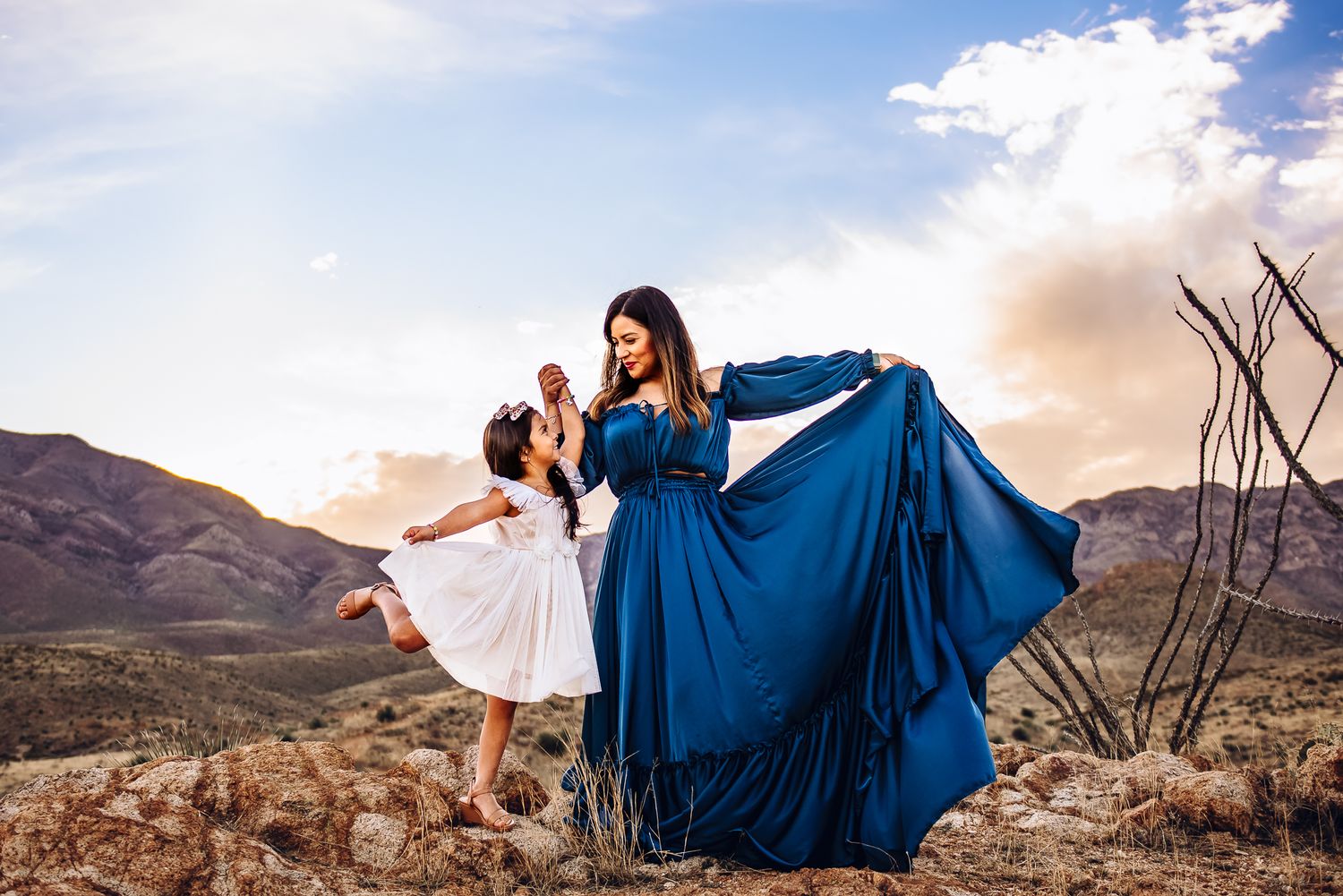 Two figures in flowing dresses dance together atop a mountain during sunset.