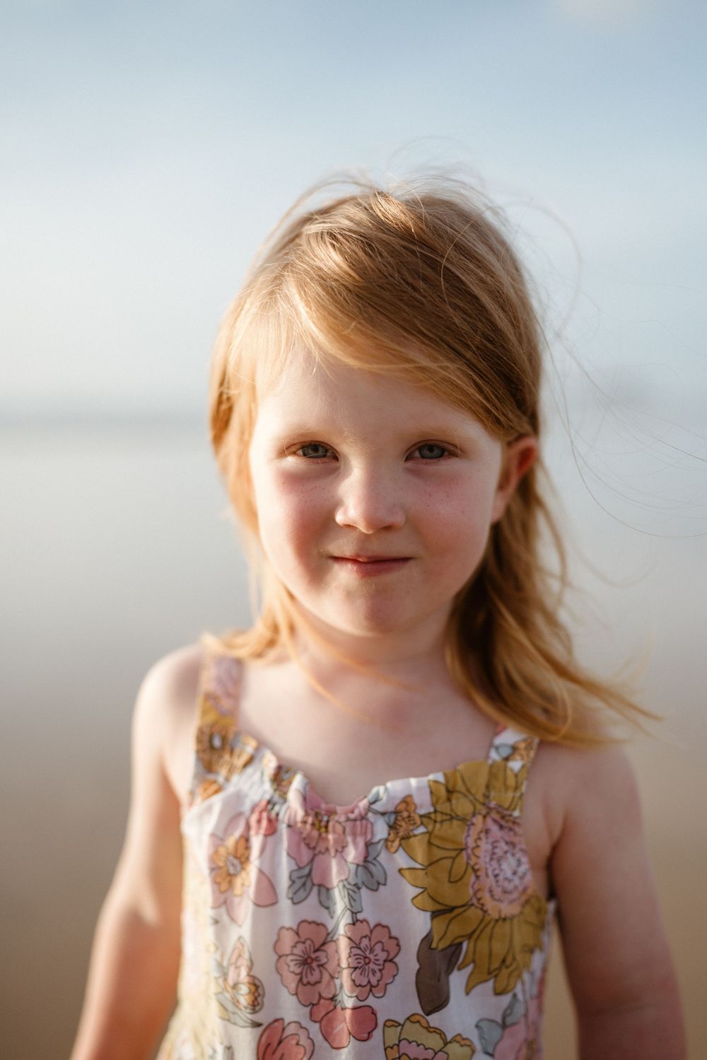Child wearing a floral sundress smiles in warm sunlight at the beach.