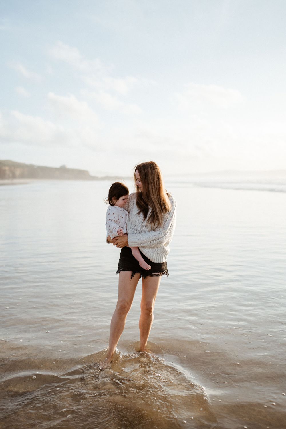 Figure standing on rocks at the beach holding a small child against a serene ocean backdrop.