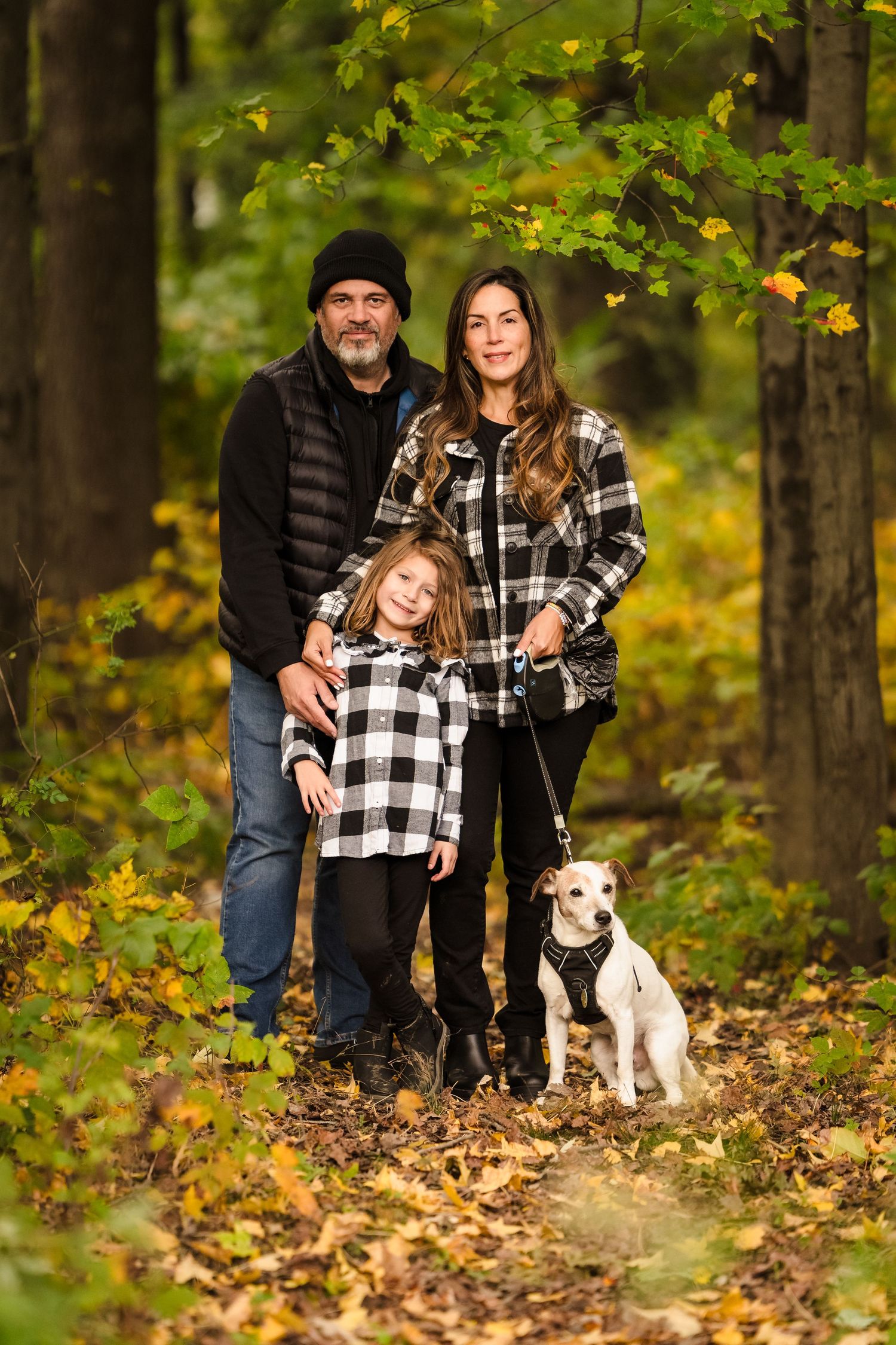 Family in matching plaid flannel poses with their white dog on autumn forest trail.