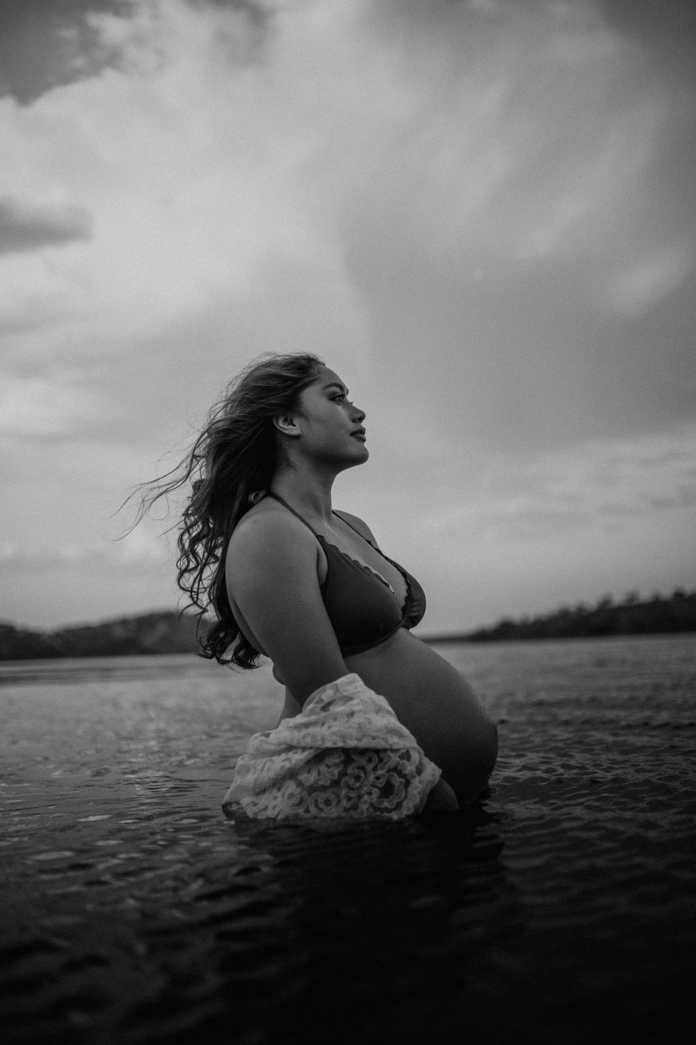 Black and white image of mama to be embracing belly while standing in water at Lake Moondarra, Mount Isa