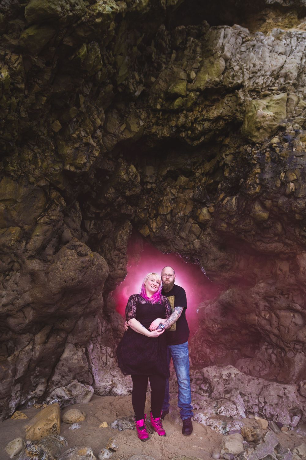 A couple stands in a cave entrance with a pink backlit glow behind them.