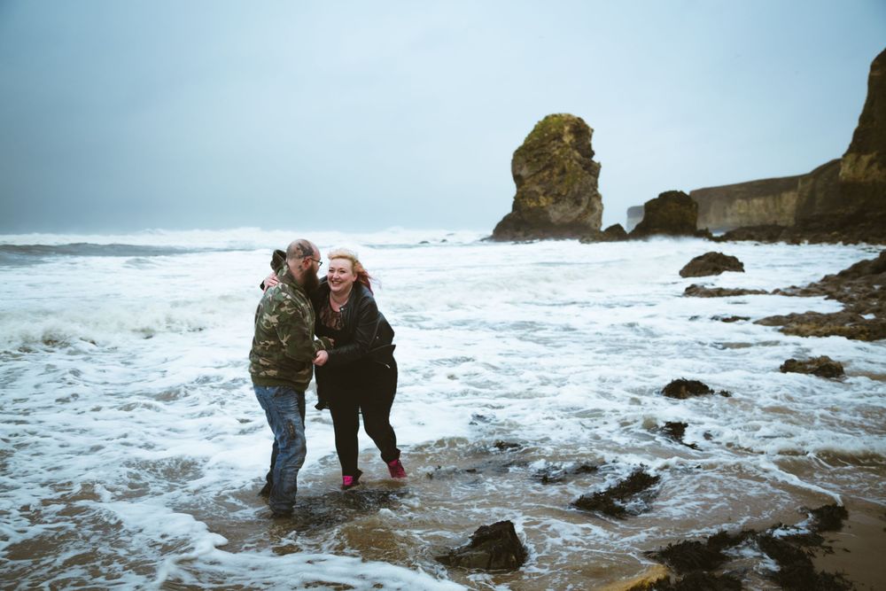 Couple embracing on rocky beach with dramatic ocean cliffs in background.