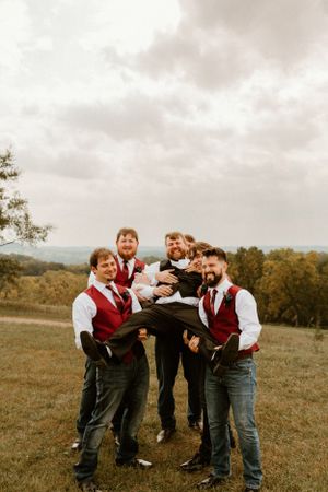 Groomsmen in red vests playfully lift someone on a hilltop under cloudy skies.