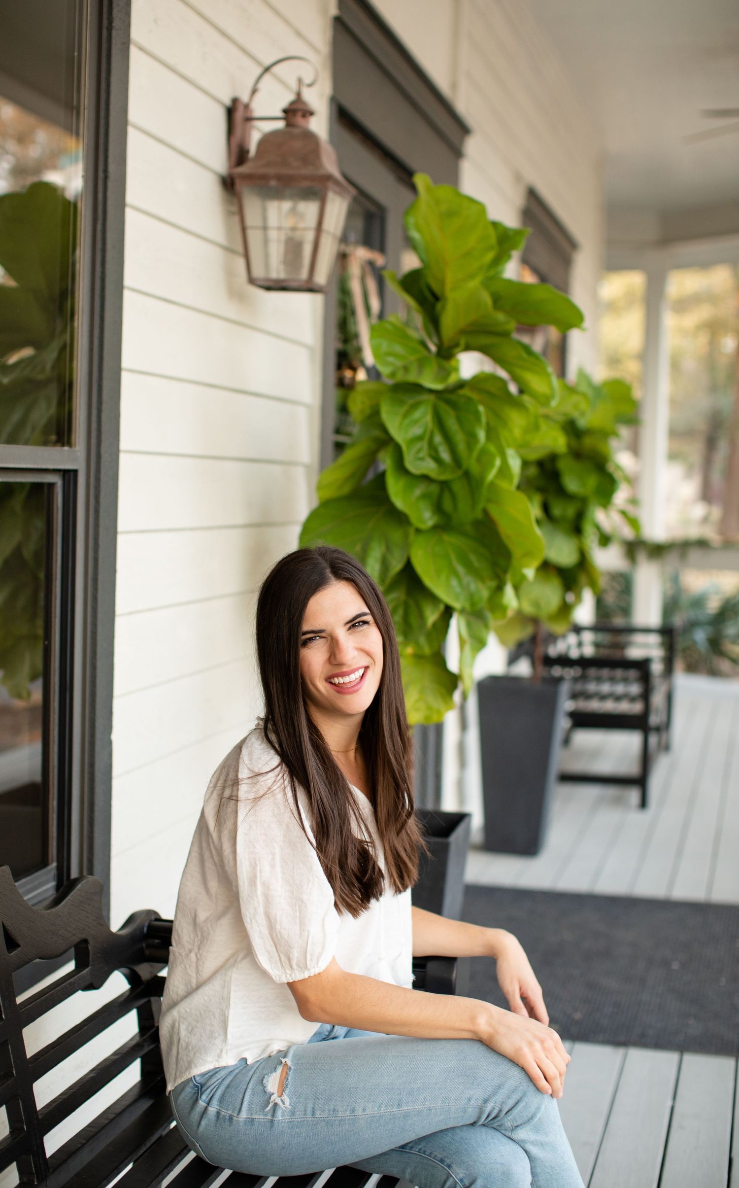 Woman in white blouse sits on black bench on front porch with fiddle leaf fig plant and copper lantern.