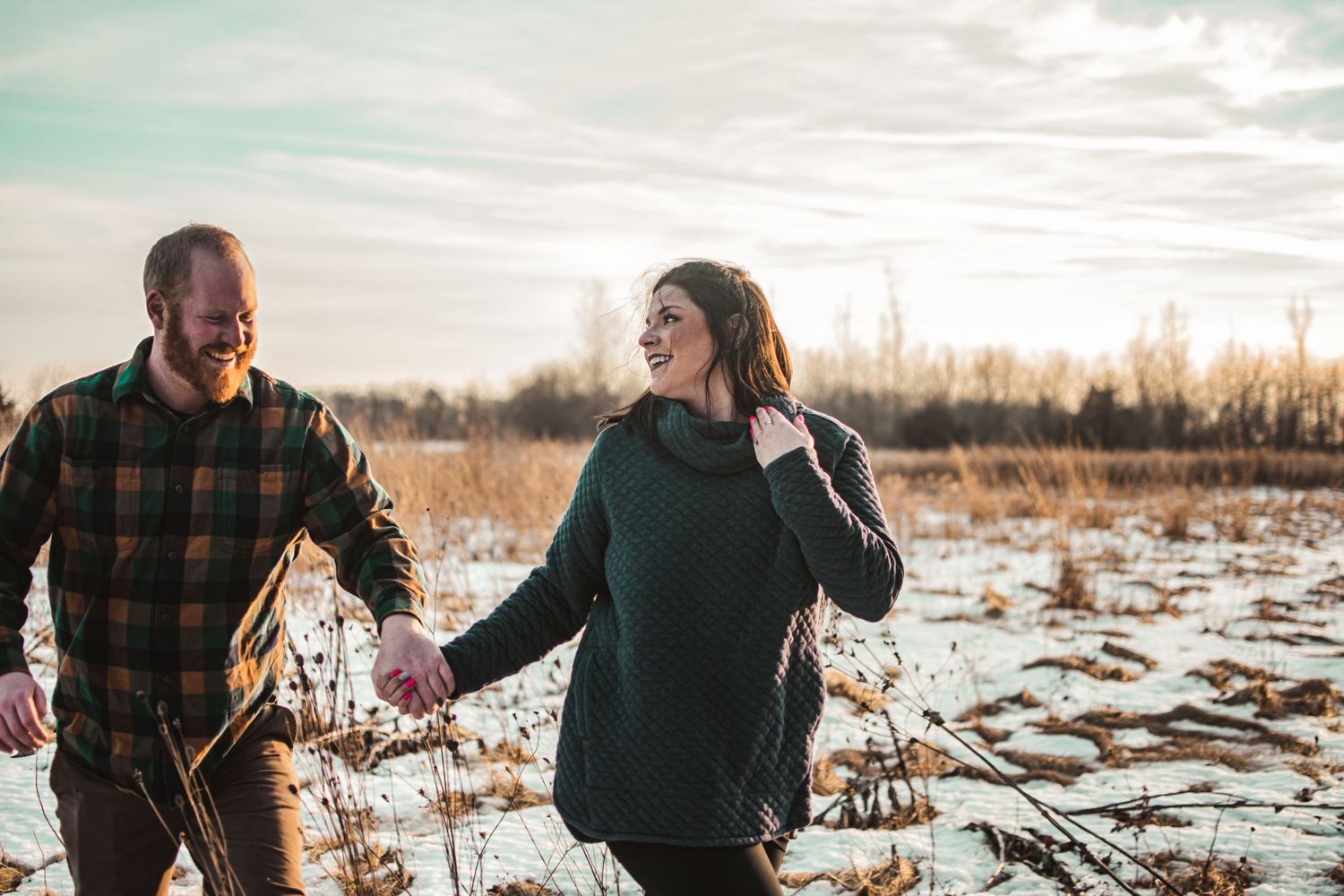 Couple walking and laughing together through snowy field at sunset.