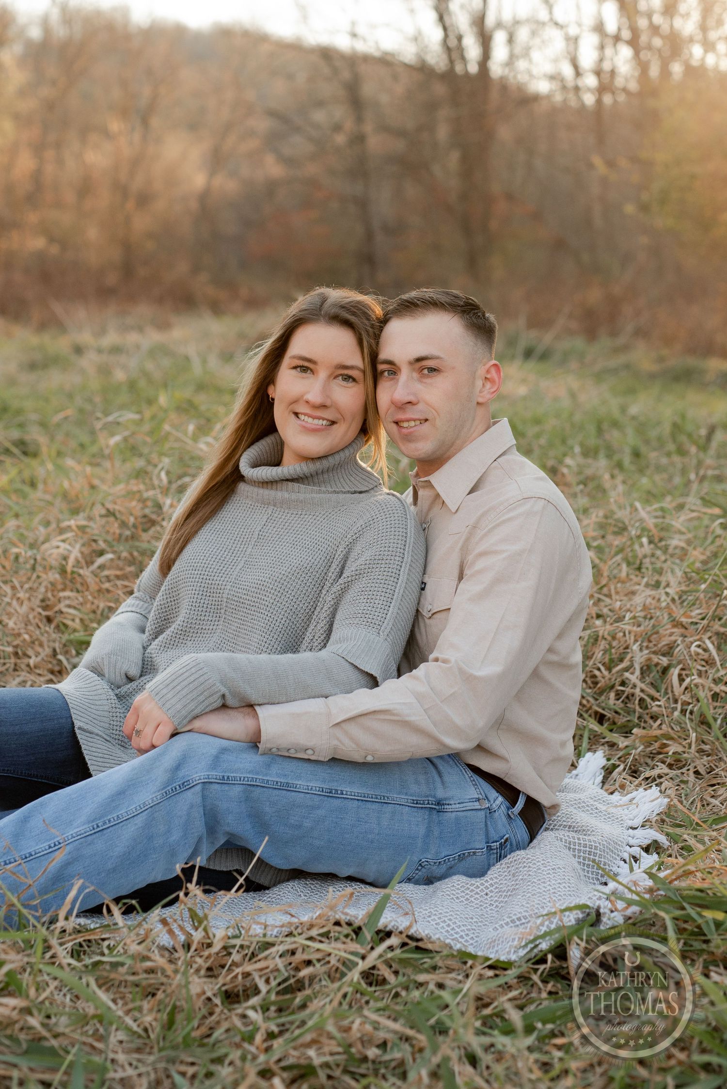 A couple sits together on grass during a fall photoshoot wearing coordinating gray and beige outfits.