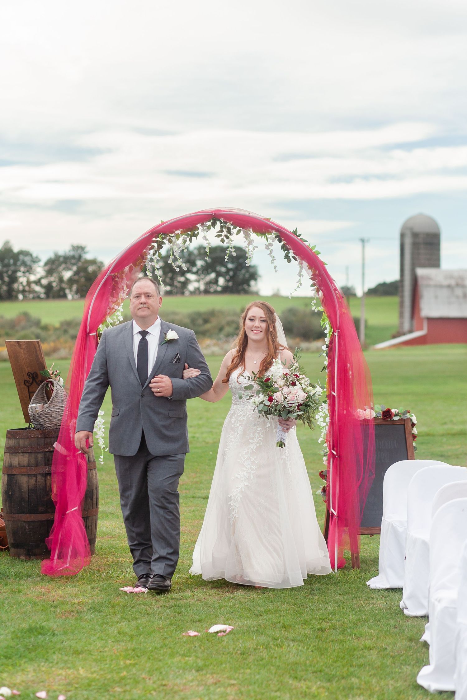 Wedding ceremony setup in a rustic farm setting with pink arch and white chairs.