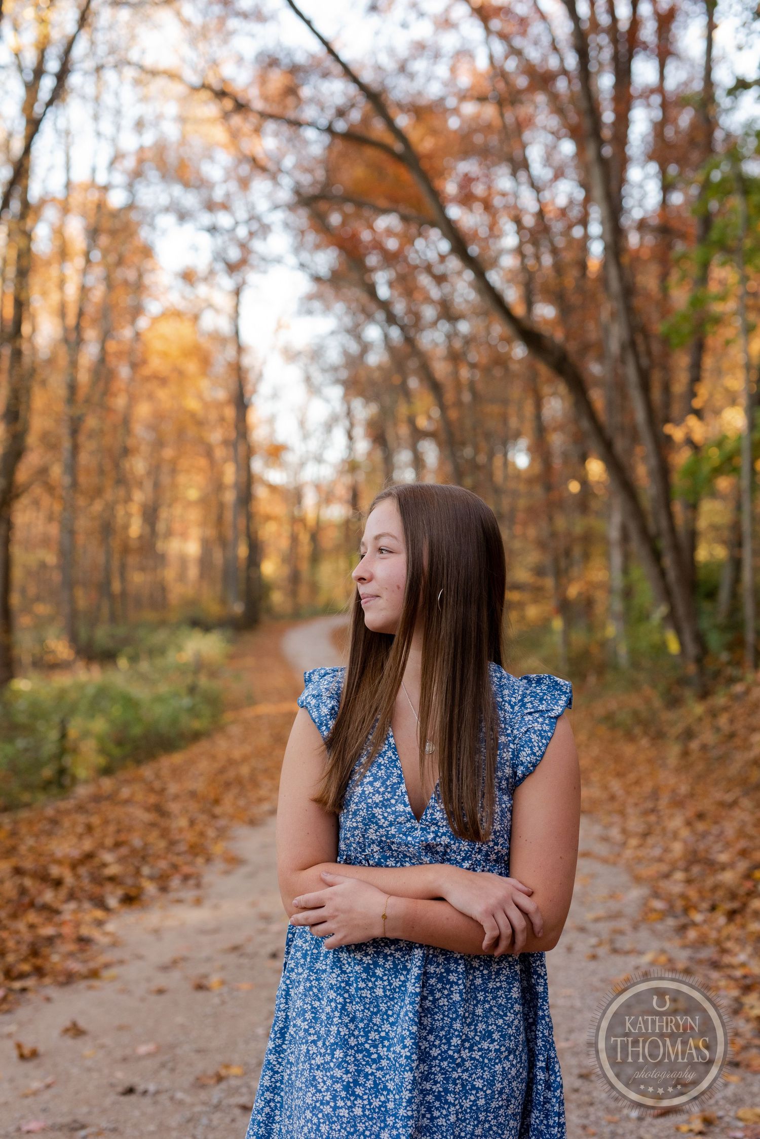 A person in blue dress poses contemplatively on an autumn trail surrounded by fall foliage.