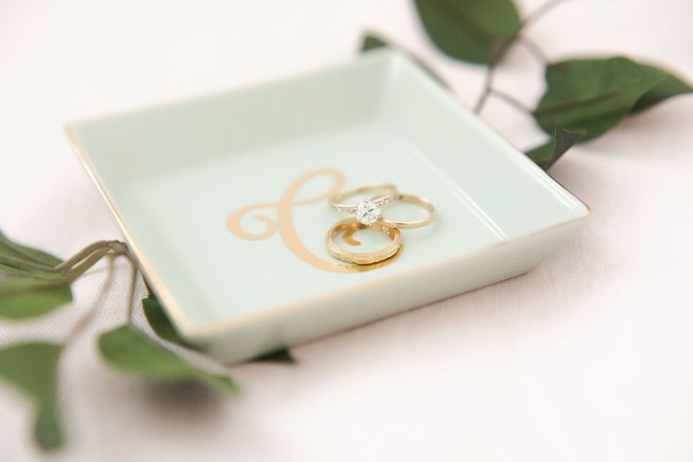 Gold diamond engagement ring and wedding bands displayed in a mint green monogram ring dish with greenery.