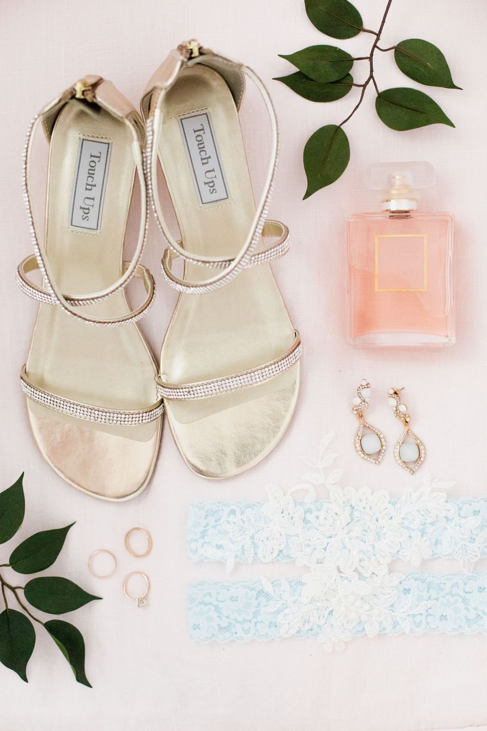 Bridal flat lay with gold sandals, blue garter, drop earrings, rings, and pink perfume bottle.