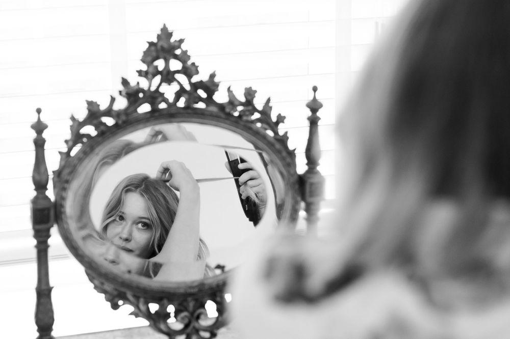 Black and white photo of a blonde woman applying makeup in an ornate vintage mirror.