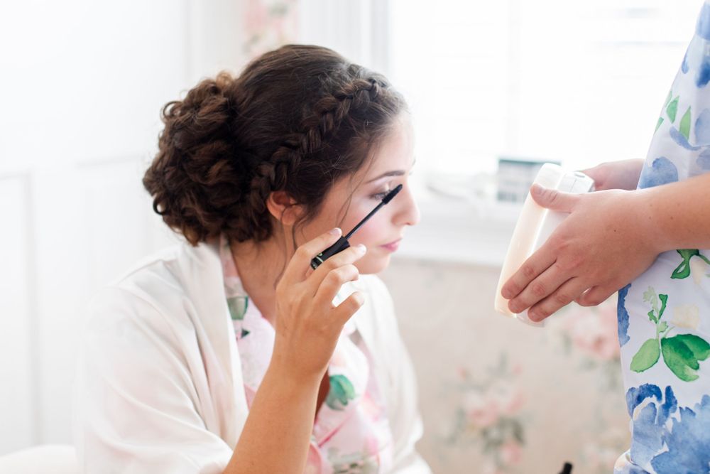 Bride in white robe applying mascara while getting ready with a makeup artist nearby.