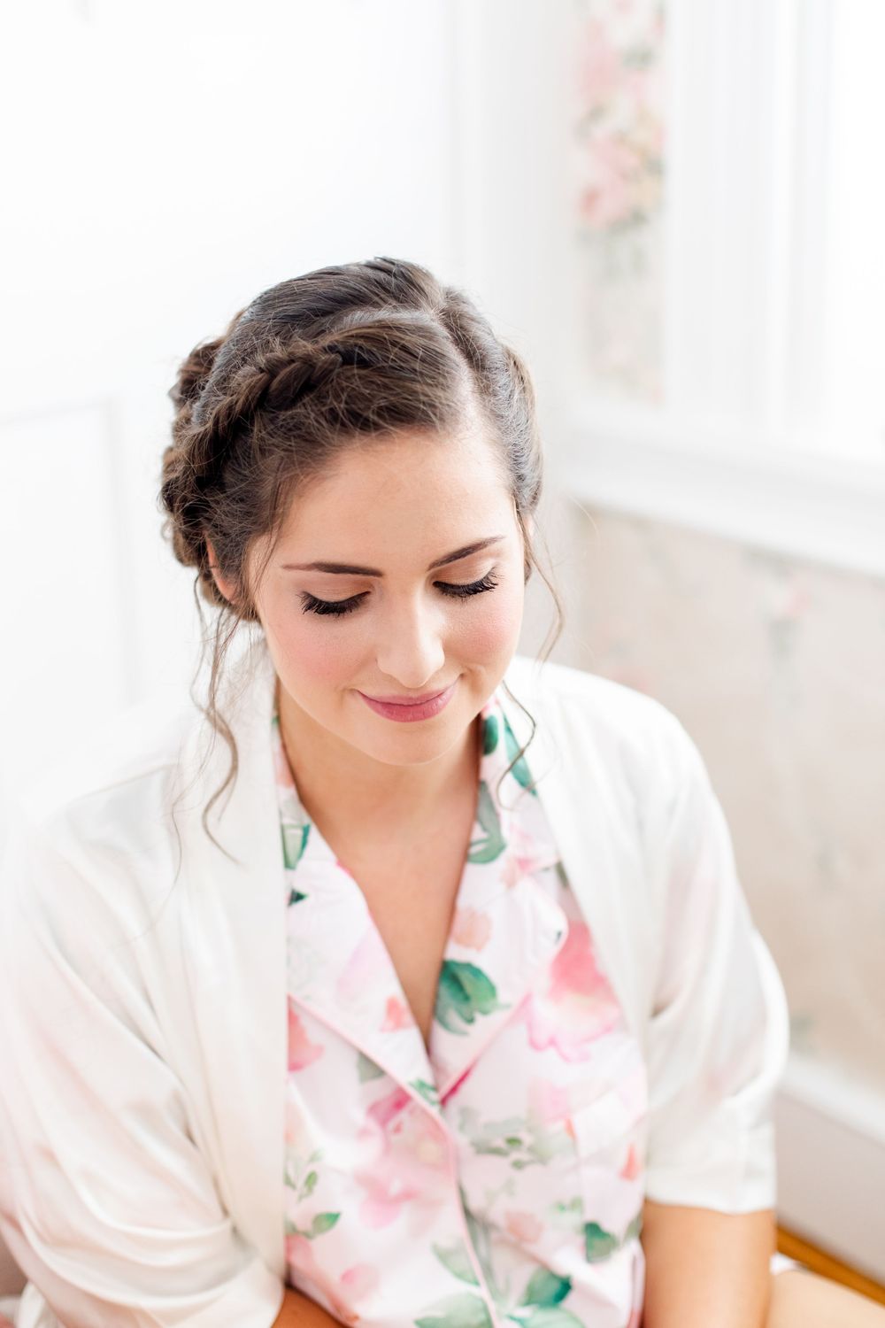 Bride with braided updo smiles down wearing floral robe during wedding morning preparations.