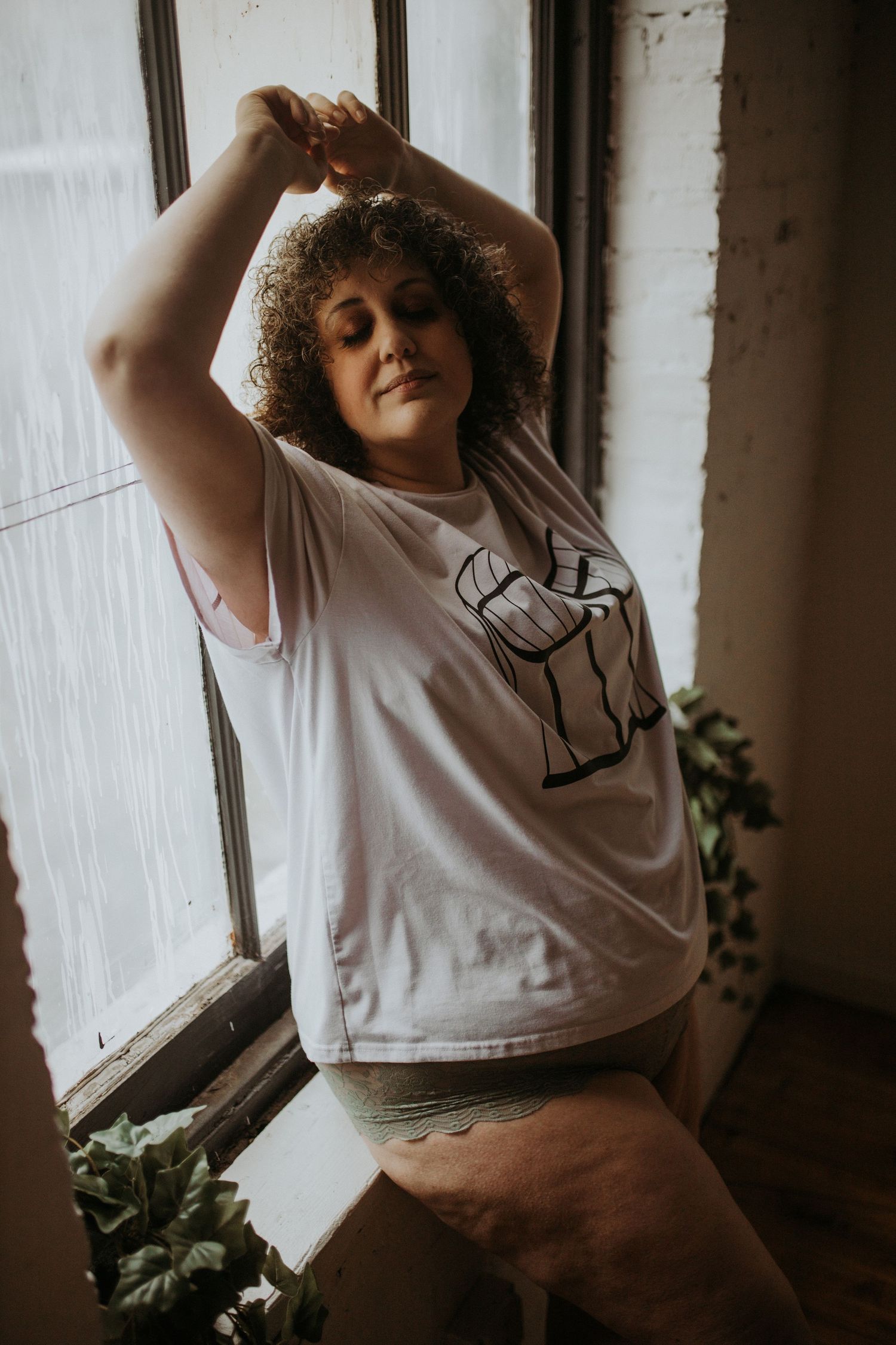 A curly-haired woman in a white t-shirt poses by a window with houseplants, creating a moody indoor portrait.