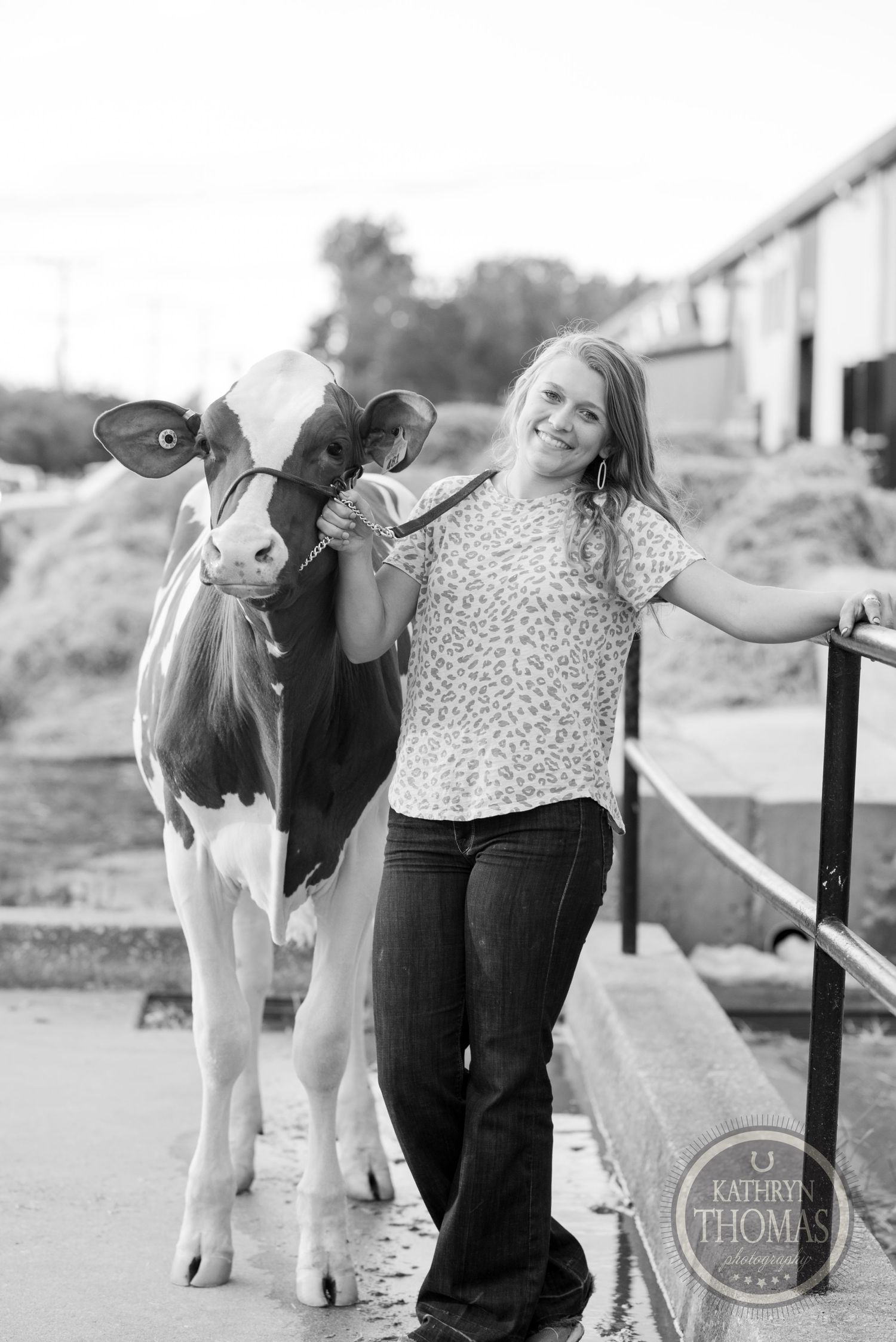 A black and white photo captures a dairy cow and person posing together by farm buildings.